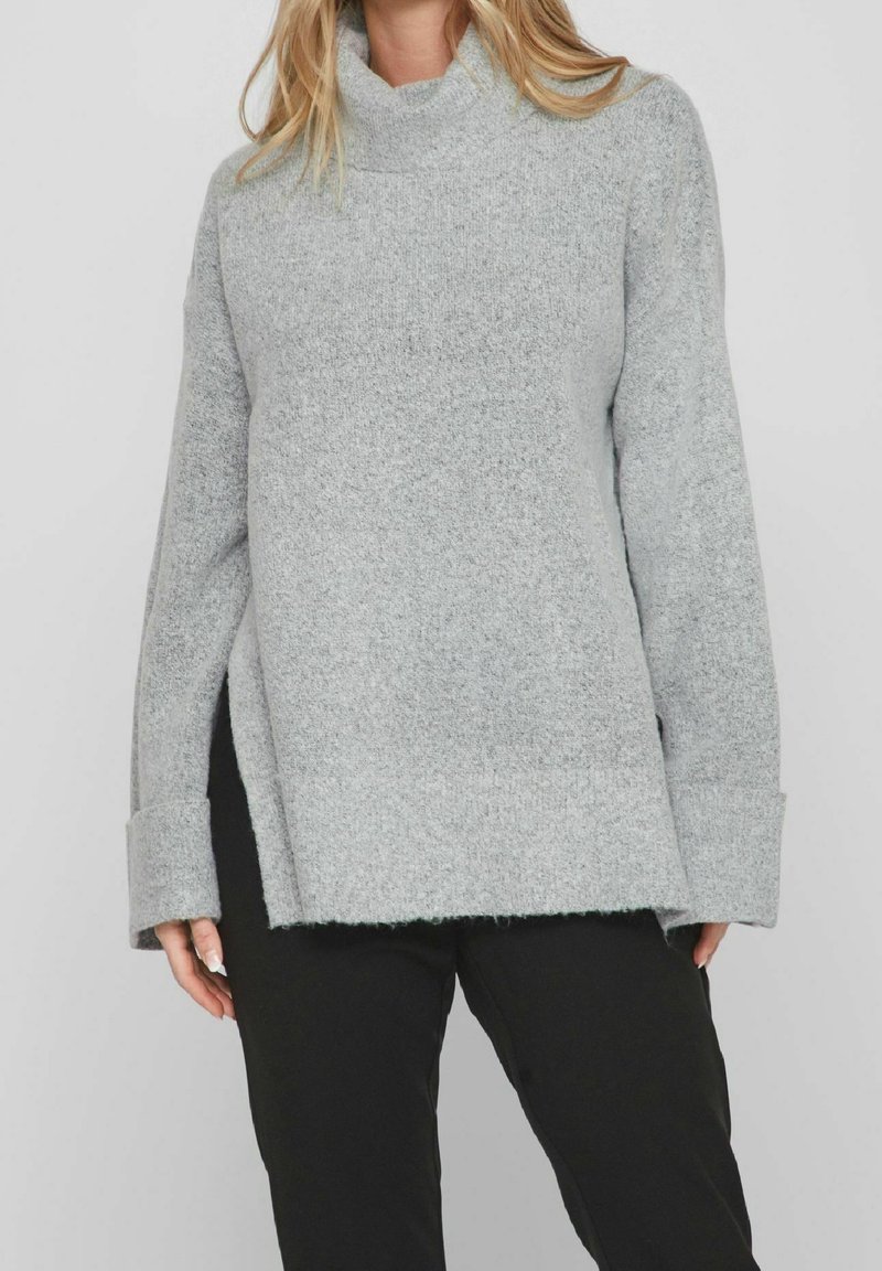 Hellgrauer Rollkragenpullover aus weichem, strukturiertem Material, mit lockerem Schnitt, seitlichen Schlitzen und breiten Bündchen. Getragen mit schwarzen Hosen.
