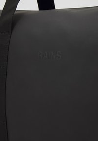 Sac de sport noir imperméable avec finition texturée, arborant un logo en relief sur le côté et de solides poignées en webbing noir.