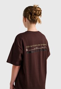 Person med flätad knut frisyr som bär en mörkbrun t-shirt med gul och vit text på baksidan om hög höjd och träning.