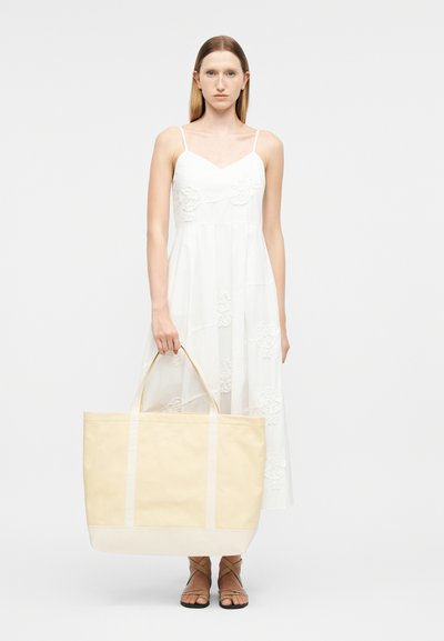 Femme en robe blanche sans manches tenant un grand sac cabas beige, portant des sandales marron à brides, debout devant un fond clair uni.