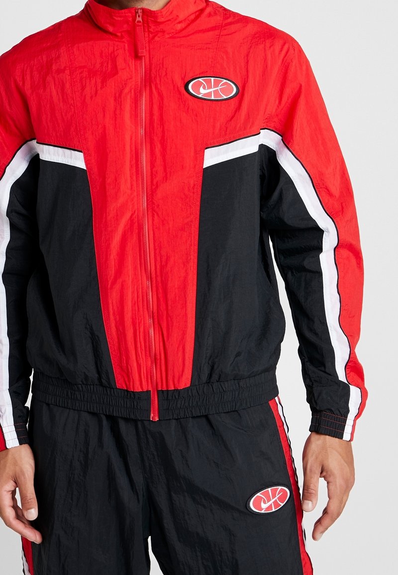 Homme portant un survêtement de sport rouge et noir avec des bandes blanches et un logo de basket-ball sur la poitrine et le short.