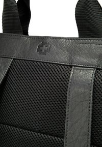 La bolsa de malla gris presenta acentos de cuero texturizado y un logotipo prominente. El diseño incluye secciones superpuestas y asas resistentes de cinta.
