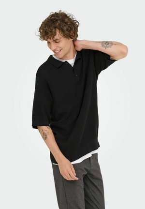 Black knitted polo shirt with a collar, short sleeves, and a relaxed fit, worn over a white shirt. Detailed arm tattoo visible.
