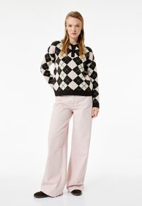 Czarny i różowy sweter w wzór argyle, z okrągłym dekoltem i czarnymi akcentami, w połączeniu z jasnoróżowymi spodniami o szerokich nogawkach i ciemnymi butami.