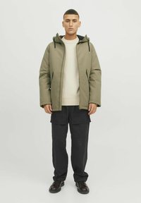 Veste matelassée vert olive avec une capuche, assortie à un pull en tricot crème et un pantalon cargo noir. Des bottes noires à lacets complètent le look.