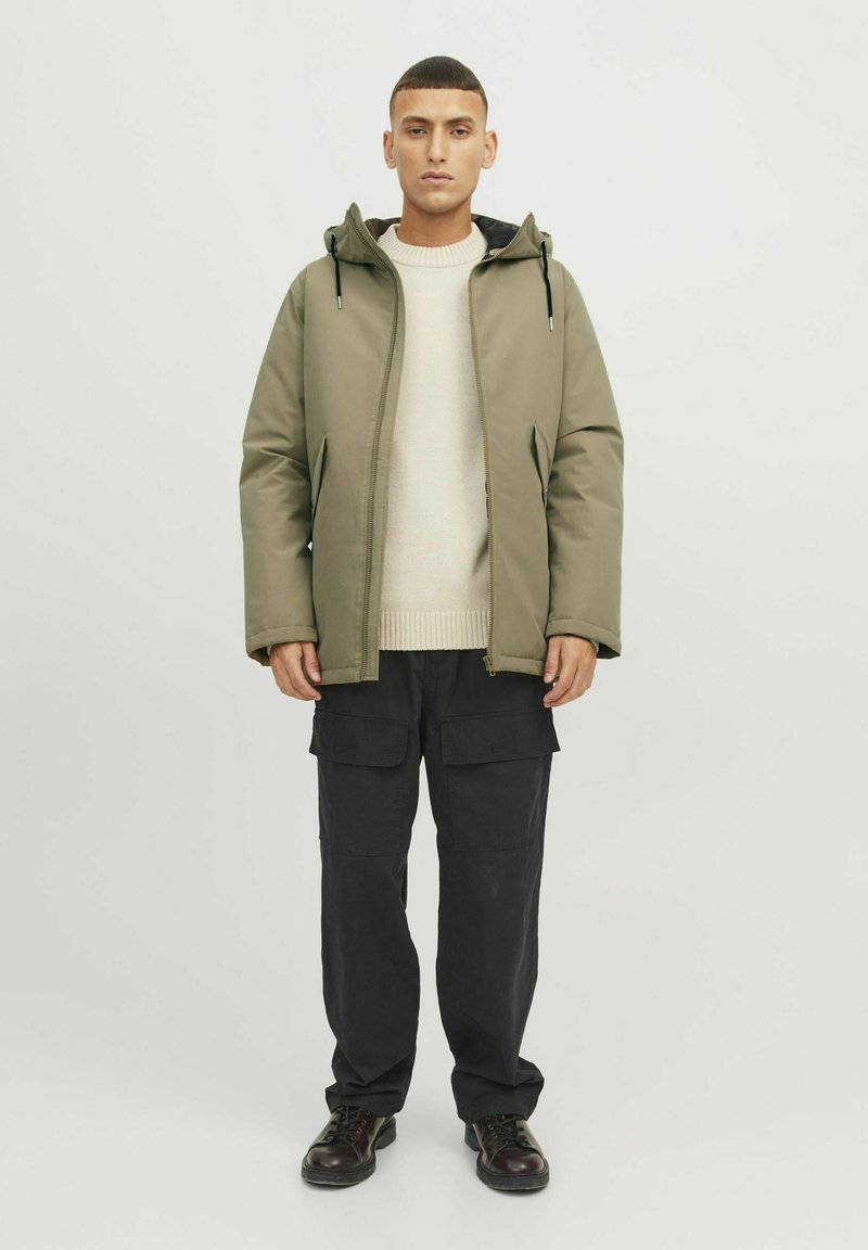 Veste matelassée vert olive avec une capuche, assortie à un pull en tricot crème et un pantalon cargo noir. Des bottes noires à lacets complètent le look.