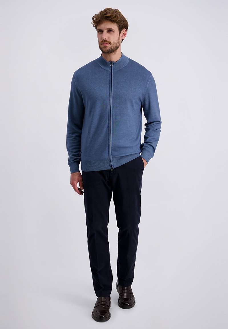 Uomo con barba che indossa un maglione blu con zip, pantaloni scuri e scarpe marroni, in piedi su uno sfondo bianco uniforme.