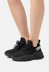 Zapatillas deportivas negras con parte superior de malla, cierre de cordones en la parte frontal y suela de goma gruesa que presenta un logotipo de marca prominente en el costado.