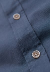 Gros plan d'un tissu bleu marine avec des boutons blancs cousus sur une patte de chemise montrant les détails des coutures.
