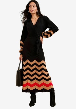 Schwarzes Wickelkleid mit langen Ärmeln, mit einem Chevron-Muster in Beige, Schwarz und Rot am Saum. Akzentuiert mit einem Bindeband in der Taille.