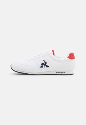 Baskets basses blanches avec languette de talon rouge, logo noir et texte « le coq sportif » sur la semelle latérale, présentées sur un fond uni.