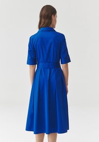 Blue dress with a collar, short sleeves, and a belt. Features a flared skirt with pleats, made of smooth fabric. Back view is shown.