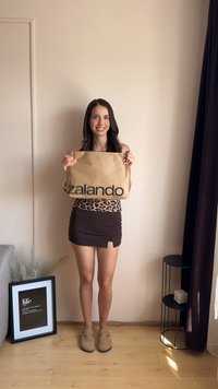 Zalando bruine papieren tas met zwart logo, vastgehouden door een persoon in een leopardprint top en een bruin minirok, staande op een houten vloer.