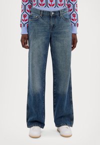 Los vaqueros de mezclilla azul claro presentan un diseño de pierna ancha, textura desteñida y bolsillos delanteros, combinados con zapatillas blancas.