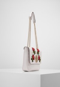 Prošívaná světle růžová kabelka se vyšívanými červenými růžemi a zelenými listy, zlatými doplňky a řetízkovým popruhem. Na přední straně ji zdobí akcentové cvočky.