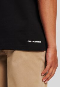 Juodas medvilninis marškinėliai su prailginta apykakle ir baltu "KARL LAGERFELD" logotipu netoli apvalkalo. Suderinta su smėlio spalvos kelnėmis, matoma ranka, remiasi į jas.