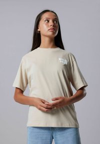 Junge Frau mit langen, dunklen Haaren, die ein beiges T-Shirt mit dem Aufdruck "Willed Away" und hellblauen Jeans trägt. Sie schaut nach oben und leicht nach rechts, mit gefalteten Händen.