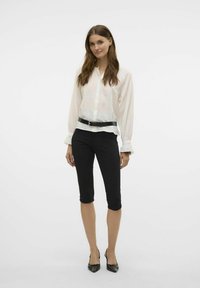 Chemise blanche à manches longues, associée à un pantalon noir court et des chaussures à talons hauts noires. La chemise présente un motif texturé.