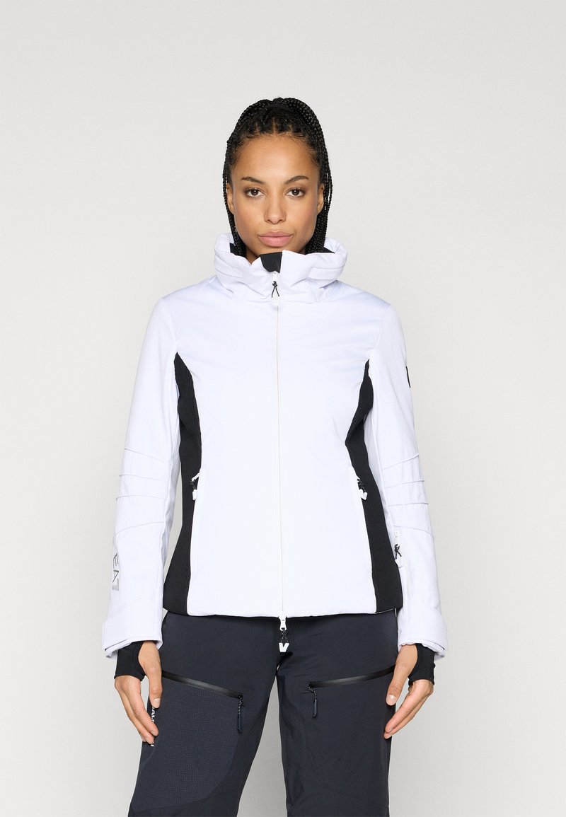 Λευκό σακάκι σκι με μαύρες πλευρικές πάνελ, ψηλό γιακά, μπροστινό φερμουάρ και φερμουάρ στις τσέπες. Μαλακή υφή με λεπτομέρειες ραφής στα μανίκια.