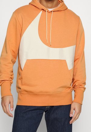 Man die een oranje hoodie draagt met een groot wit swoosh-ontwerp en donkerblauwe spijkerbroek, staand met ontspannen armen.