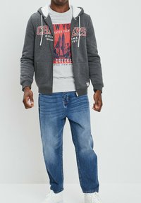 Sudadera gris con cremallera y cordones blancos, con bolsillo; camiseta gráfica gris con estampado rojo; vaqueros azules; zapatillas blancas.