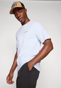 Camiseta blanca de manga corta con un logo "Patagonia" en azul claro, combinada con pantalones negros, llevada debajo de una gorra trucker en tonos marrones y negros.