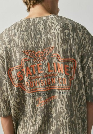 Person trägt ein graues und beiges Hemd mit Camouflage-Muster, orangefarbenem "State Line Provision Co" Text und einer Adler-Grafik auf dem Rücken.