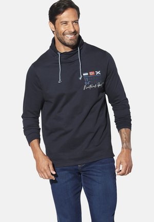 Navy blue sweatshirt with a high collar, drawstrings, and logo details in white and colored flags. Worn with blue jeans.