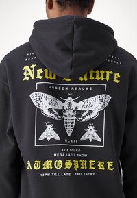 Felpa nera con cappuccio caratterizzata da una grande grafica bianca e gialla di falene e dettagli dell'evento. Il materiale è morbido, con una vestibilità ampia e cappuccio regolabile.