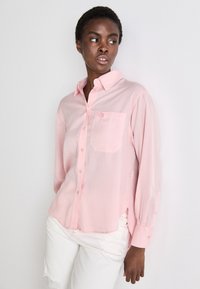 Camicia a maniche lunghe rosa chiaro con colletto a bottone, taschino sul petto e orlo leggermente arrotondato. Il tessuto appare liscio e leggero.