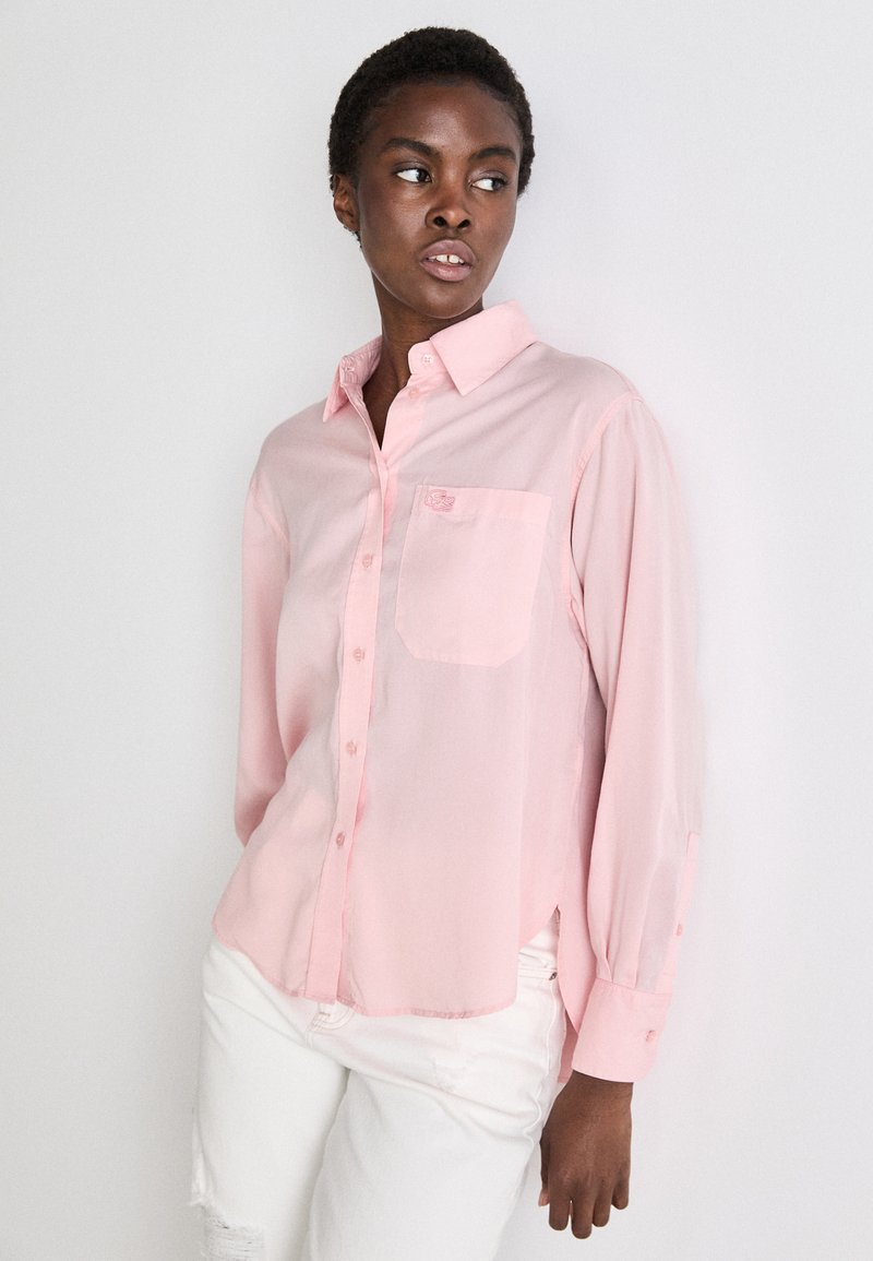 Chemise à manches longues rose clair avec un col boutonné, une poche poitrine et un ourlet légèrement arrondi. Le tissu semble lisse et léger.