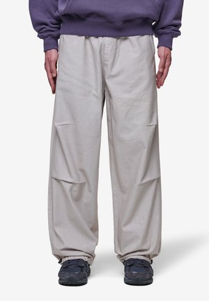 Személy laza, világos bézs színű cargo nadrágot visel, amelynek elasztikus mandzsettái vannak, és sötétkék sportcipőt párosít egy lila pulóverrel.