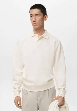 Young man wearing a cream long-sleeve polo shirt and beige trousers, holding a folded cream sweater against a plain white background.