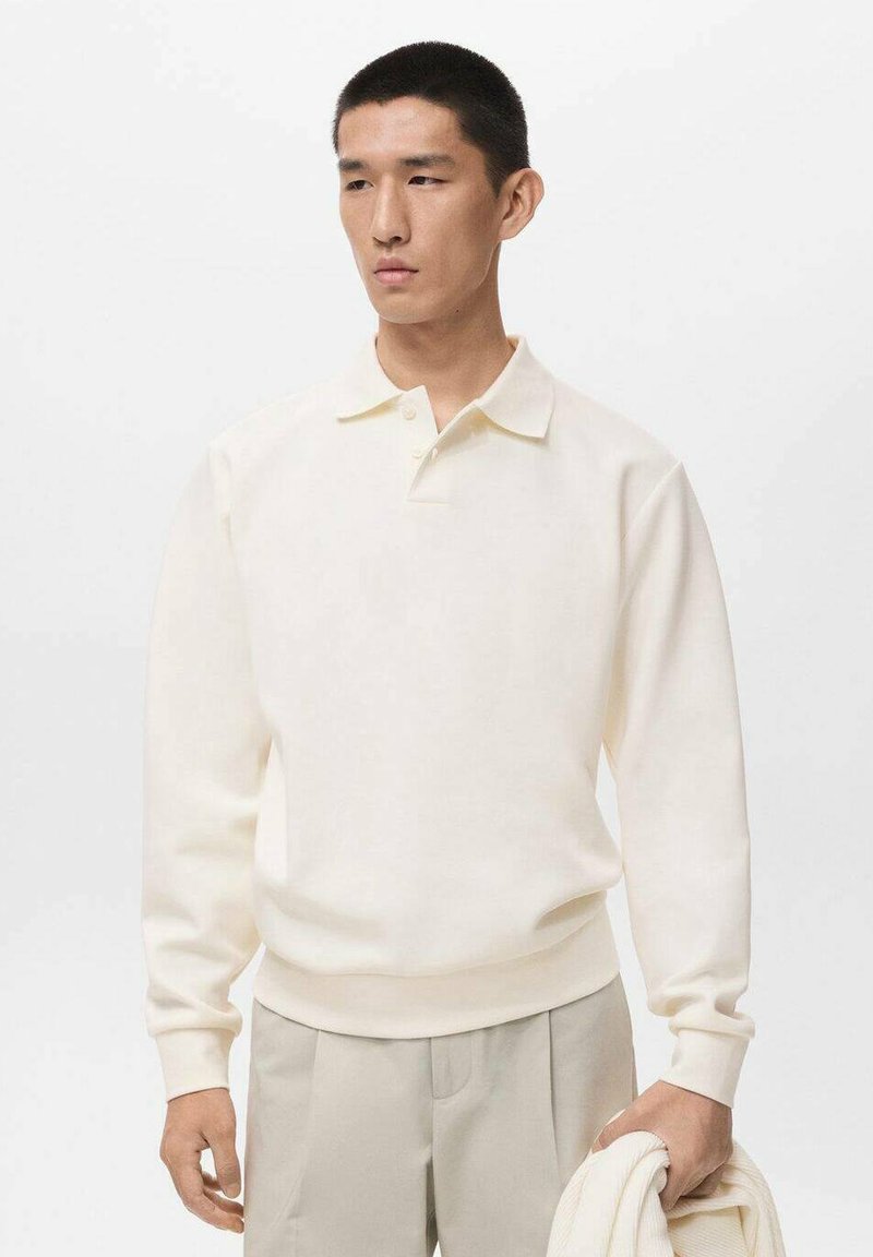 Junger Mann in einem cremefarbenen Langarm-Polohemd und beigen Hosen, hält einen gefalteten cremefarbenen Pullover vor einem schlichten weißen Hintergrund.