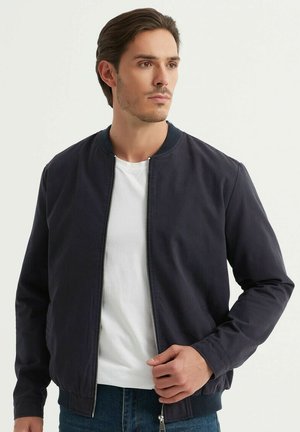 Veste bombardier marine avec une texture lisse, col côtelé, fermeture éclair à l'avant et poches latérales, portée sur un t-shirt à col rond blanc.
