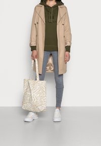 Abrigo trench beige, sudadera con capucha verde, vaqueros grises, zapatillas blancas, sosteniendo un bolso tote beige con patrón. Tela suave y diseño casual.