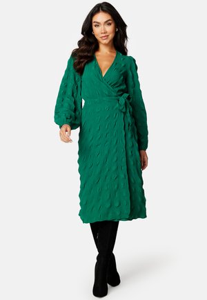 Groene wikkeljurk van gestructureerd stof, met lange pofmouwen en een geknoopte taille. Gestyled met zwarte kniehoge laarzen.