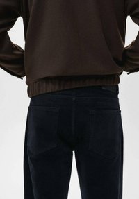 Jersey marrón oscuro con dobladillo acanalado, combinado con pantalones de pana azul oscuro que tienen bolsillos traseros y una superficie texturizada.