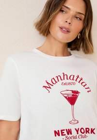 Bílý bavlněný tričko s červeným grafickým potiskem koktejlové sklenice a textem "Manhattan Est. 1870" a "New York Social Club." Kulatý výstřih.
