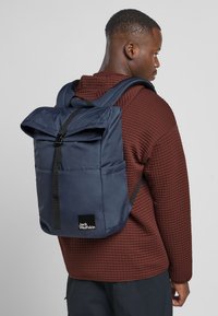 Zaino blu navy con design a roll-top, realizzato in tessuto testurizzato. Dotato di spallacci neri e logo Jack Wolfskin sul davanti.