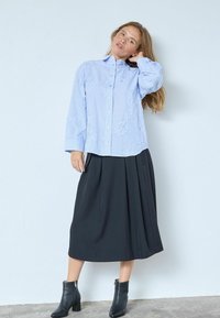 Vaaleansininen, napitettava paita, jossa on kukkakirjailu, yhdistetty mustaan laskostettuun midihameeseen ja mustiin nilkkureihin harmaalla lattialla.