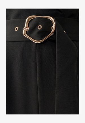 Ceinture en tissu noir avec une boucle en or de forme irrégulière, affichant une texture lisse et une boucle pour ajustement, attachée à un vêtement ajusté.