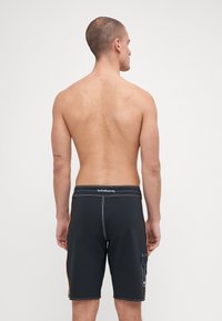 Shorts de bain noirs avec des rayures latérales orange, une taille ajustée sur mesure et une poche unique sur la jambe gauche, mettant en valeur des coutures contrastantes.