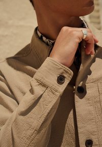 Beige Jacke aus einem strukturierten Stoff, mit geknöpften Manschetten und dunklen Knöpfen. Ein gemusterter Kragen ist unter der Jacke sichtbar.
