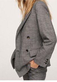 Szara dwurzędowa marynarka w kratę z teksturowanej tkaniny, z czarnymi guzikami i bocznymi kieszeniami. Dopasowany krój podkreśla kształt i strukturę.
