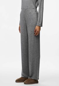 Cilvēks, kurš ģērbjas brīvos pelēkos biksēs, atbilstošā garās piedurknes krekla un brūnos slaidajos apavos, stāv pret vienkāršu baltu fona.