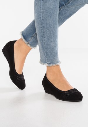 Fødder iført sorte kile-ballet-sko med nagle-accenter og lyseblå jeans med frynset kant på et glat hvidt gulv.