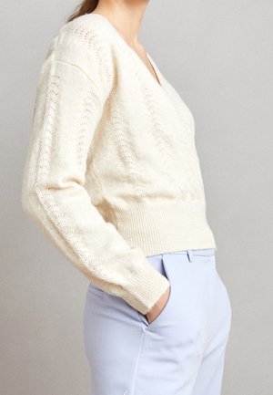 V-kaula-aukollinen kerma-vaalea neulepusero avoimella kuviolla ja resorireunuksella. Yhdistetty vaaleansinisten housujen kanssa. Pehmeä rakenne ja istuva siluetti.