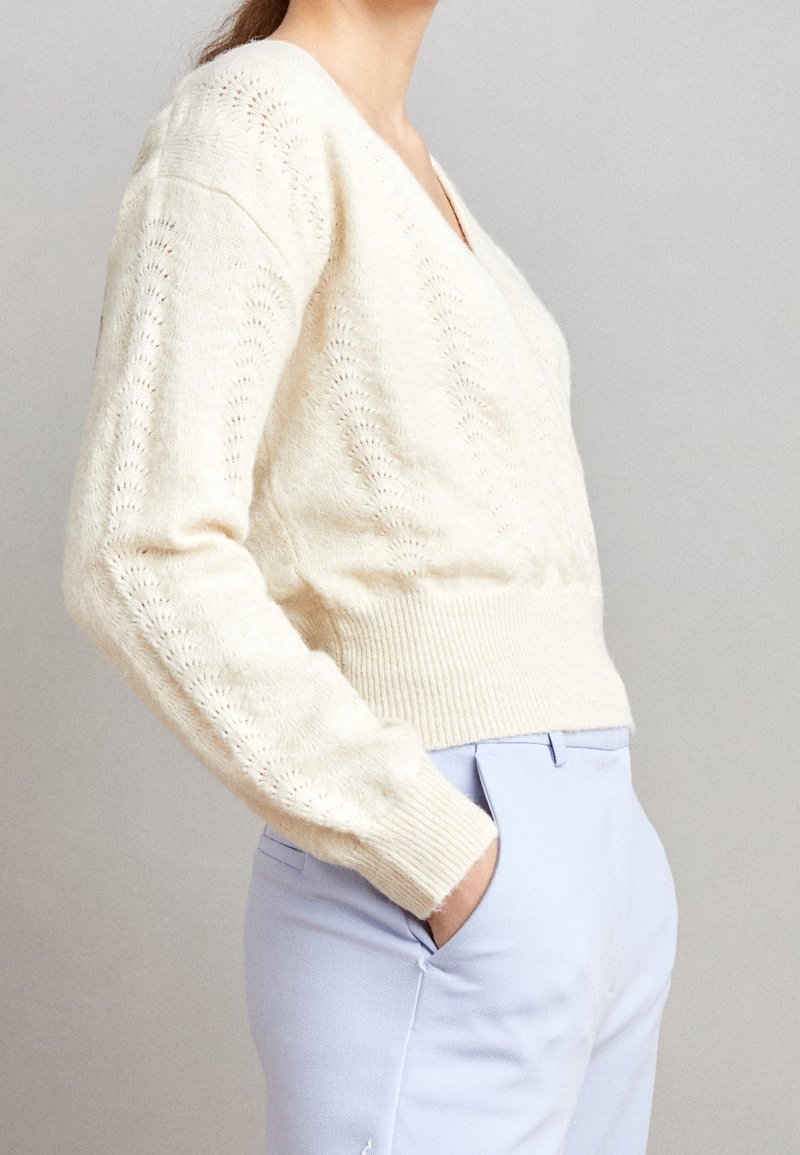 V-kaula-aukollinen kerma-vaalea neulepusero avoimella kuviolla ja resorireunuksella. Yhdistetty vaaleansinisten housujen kanssa. Pehmeä rakenne ja istuva siluetti.