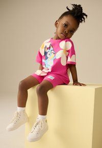 Maglietta rosa a maniche corte con personaggio dei cartoni animati, abbinata a pantaloncini coordinati. Scarpe da ginnastica bianche alte e calzini alla caviglia. Sfondo beige semplice.